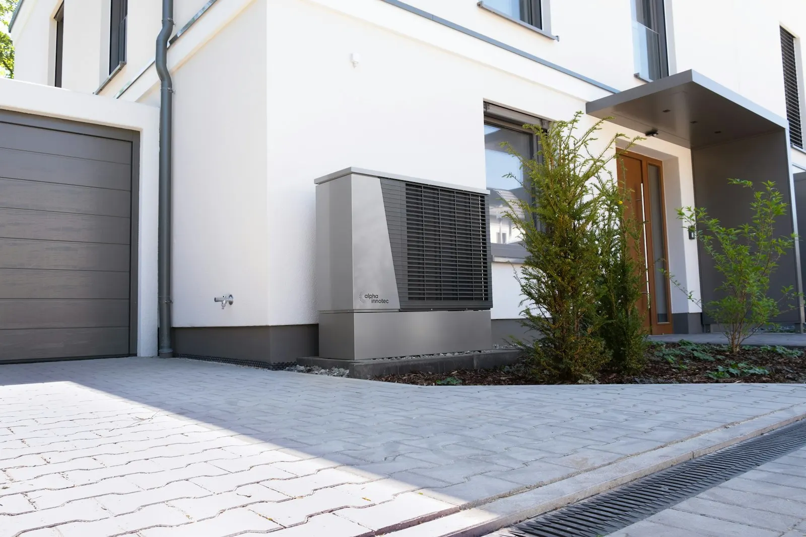 Pompe à chaleur posée contre le mur enduit blanc d'une maison, au bord d'une allée pavée