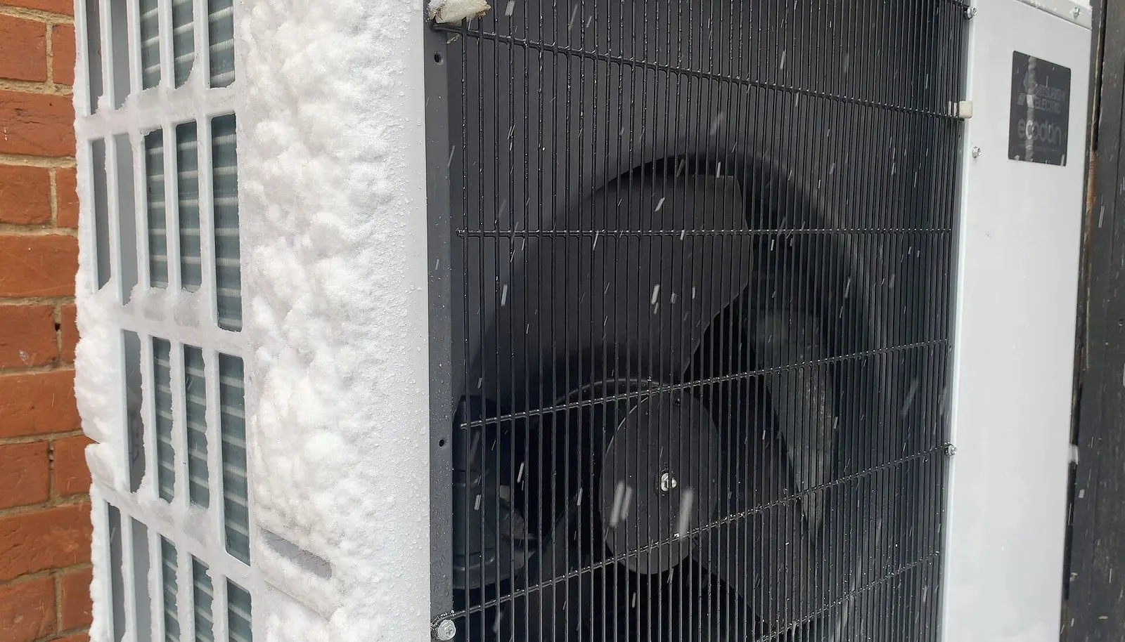 Unité extérieure d'une pompe à chaleur en marche sous la neige, contre un mur de briques enneigé